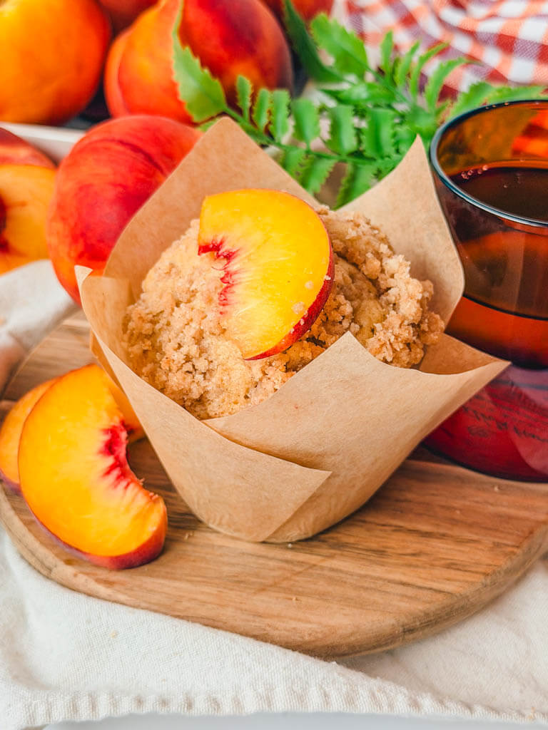 a peach muffin in a tulip liner with a fresh peach slice on top of the muffin.