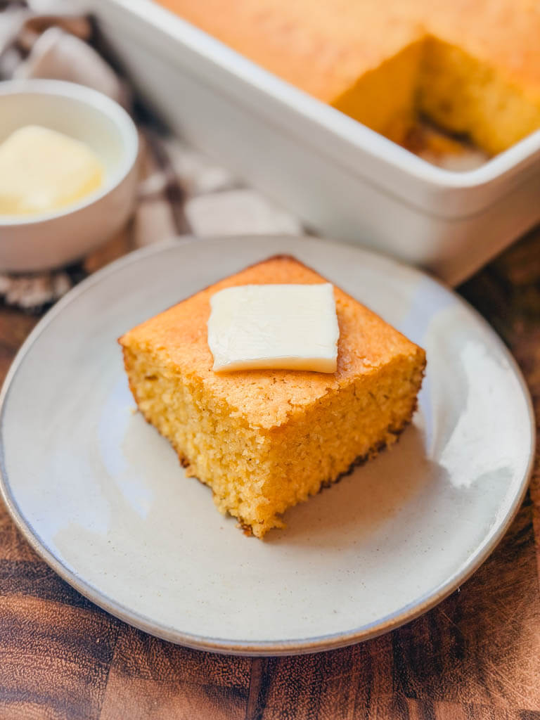 Slice of homemade sweet cornbread topped with butter on a cream colored plate.