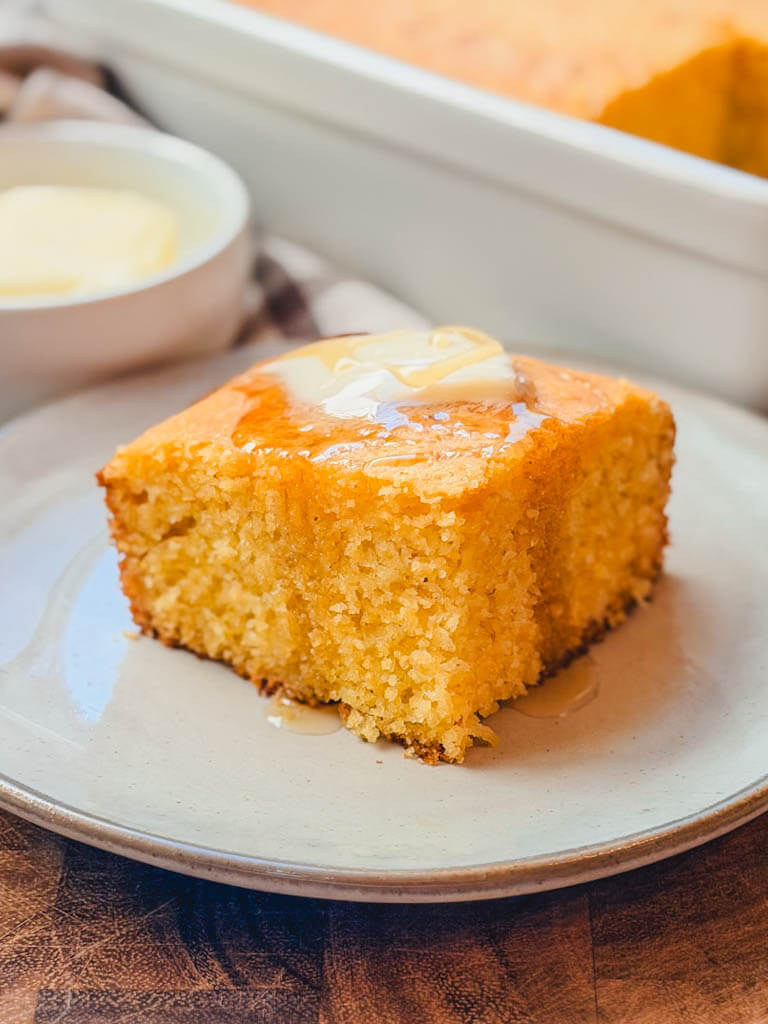 close up of a serving of cornbread on a plate with a butter bowl in the background.