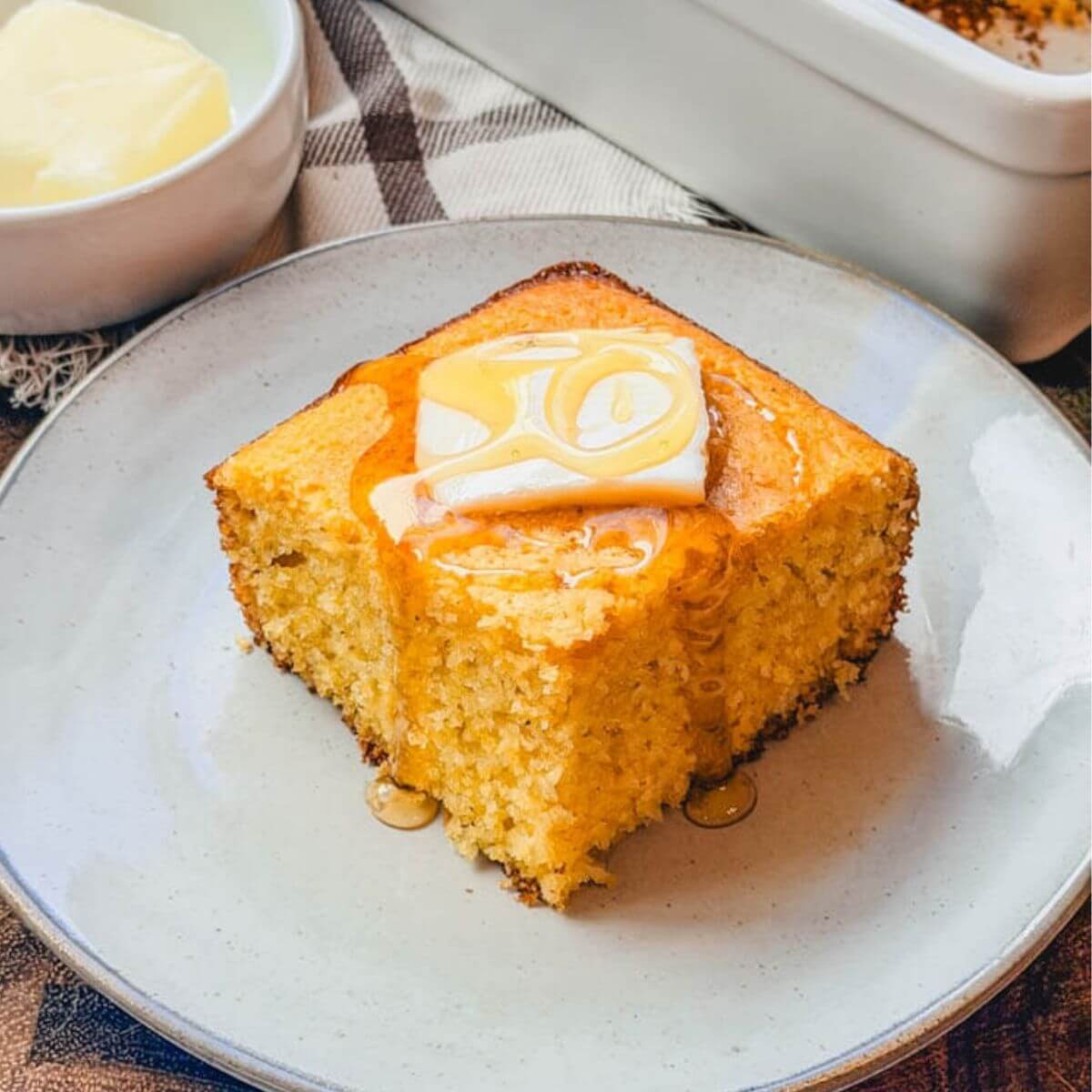 close up of a single serving of homemade sweet cornbread on a plate with a pat of butter and drizzle of honey over the top.