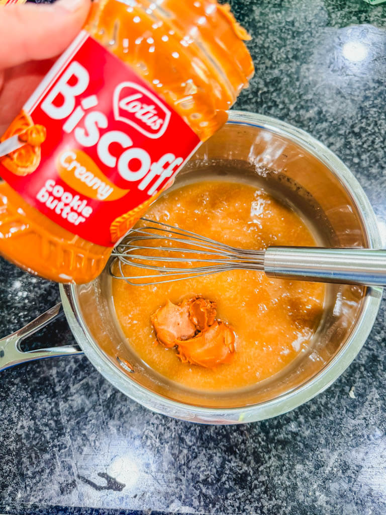 Biscoff cookie butter being added to warm brown sugar icing in a saucepan.