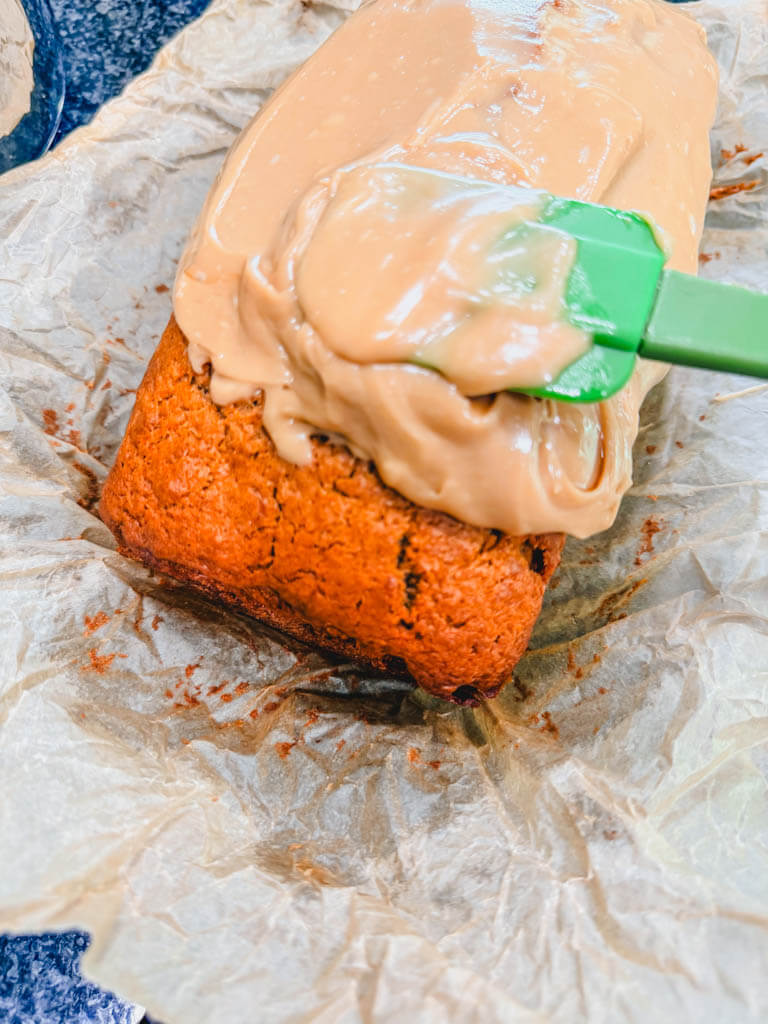 Brown sugar Biscoff icing being spread over a cooled loaf of banana bread with a spatula.
