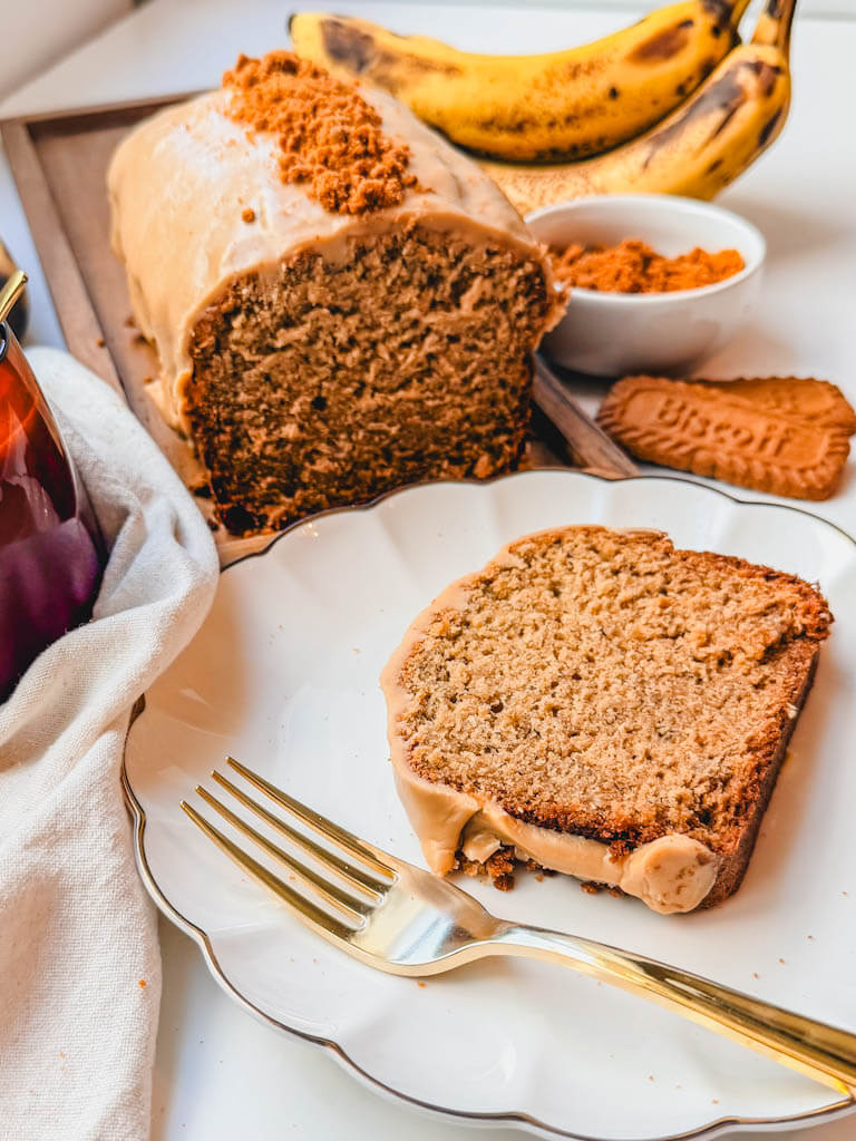 Slice of Biscoff banana bread with iced loaf, ripe bananas, and Biscoff cookie crumbs in the background.