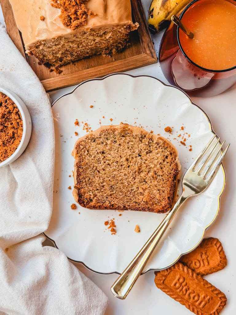 Slice of Biscoff banana bread with the iced loaf, crushed Biscoff cookies, and a drink in the background.