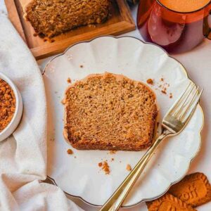 Slice of Biscoff banana bread on a white plate with Biscoff cookies, iced loaf, and drink.
