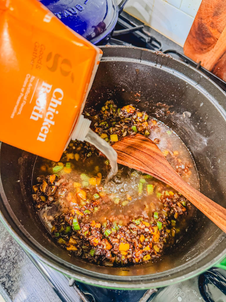 Chicken broth being poured into a Dutch oven with dark roux and vegetables.