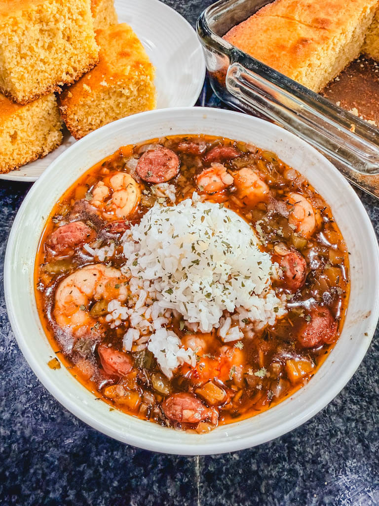 close up of shrimp and andouille sausage gumbo serving in a white bowl with rice and cornbread in the background.