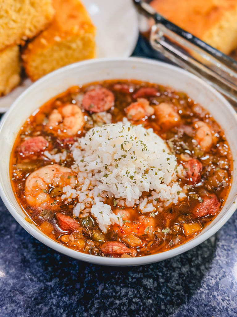 gumbo in a white bowl with rice in the center of the gumbo and cornbread next to the bowl.