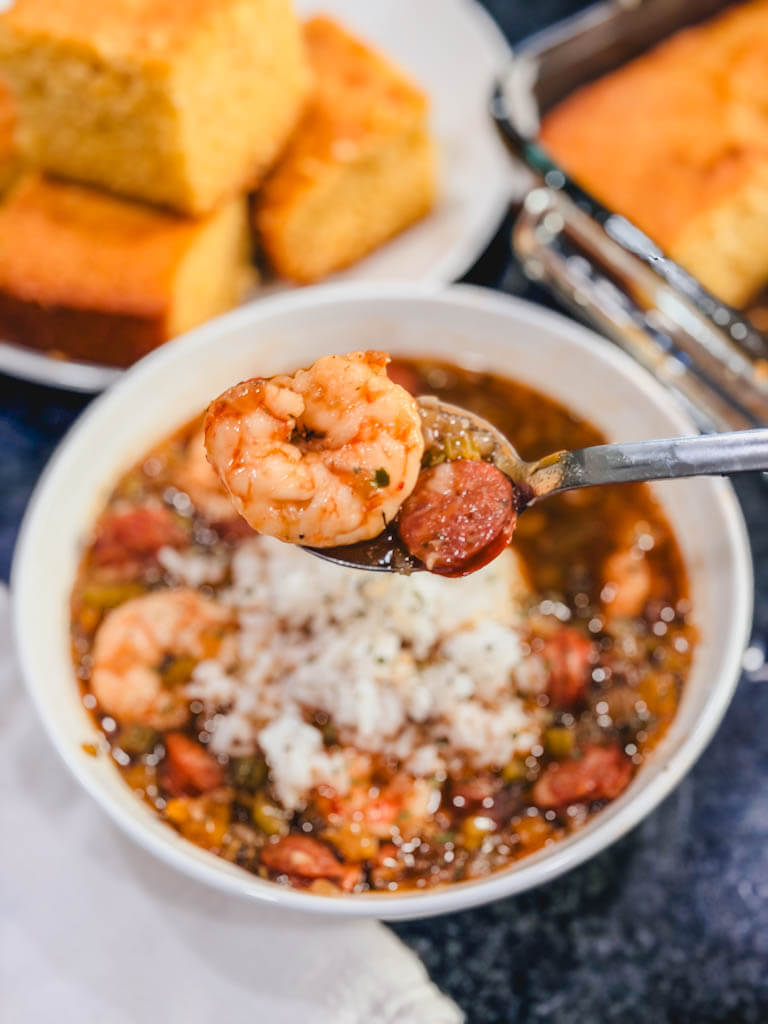 shrimp and sausage on a spoon being held over a white bowl of gumbo and rice.